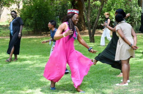 A group of women joyfully dance and interact on a grassy lawn surrounded by trees. One woman in the foreground wears a bright pink outfit and a beaded headband, smiling and mid-step, while others laugh and move freely in the background. The scene conveys a sense of celebration and connection in a natural setting.